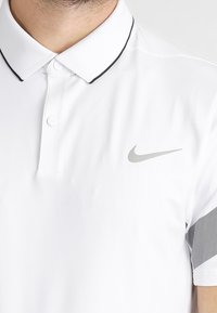Bílé polo tričko s černým lemem na límci, s kovovým logem Nike na hrudi a vzorovanými šedými akcenty na ramenou.