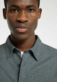 Grijze button-up shirt met een gestructureerde weefsel, met een kraag en een enkele knoop bij het nekgedeelte, tegen een neutrale achtergrond.