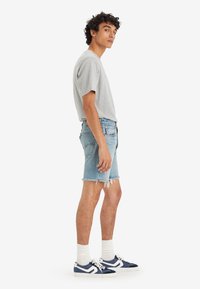 Camiseta de algodón gris, pantalones cortos de mezclilla azul claro con dobladillo deshilachado, calcetines blancos y zapatillas deportivas marinas con acentos blancos, en pose de pie.