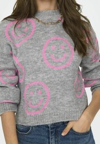 Szary sweter dzianinowy z dużymi różowymi wzorami uśmiechniętej buzi, z ribbrowymi mankietami i okrągłym dekoltem, zestawiony z niebieskimi dżinsami i złotym naszyjnikiem.