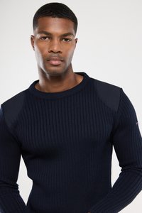 Granatowy sweter z prążkowanej dzianiny z dopasowanymi rękawami, okrągłym dekoltem i kontrastowymi łatami na ramionach. Materiał wydaje się strukturalny i przytulny.