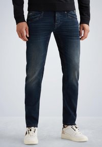 Jeans de mezclilla azul oscuro con corte recto, que presentan zonas sutilmente descoloridas, bolsillos frontales y combinados con zapatillas de color claro.