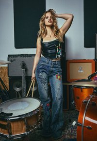 Musta nahkapaidat, joissa on monivärinen kuvio, yhdistettynä sinisiin leveälahkeisiin farkkuihin, joissa on lohikäärme- ja kukkakirjontaa. Puiset rumpukepit kädessä.