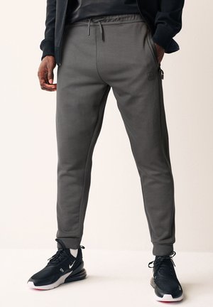 Joggers grigi con gamba affusolata, vita elasticizzata, tasche laterali e un discreto dettaglio del logo. Realizzati in un tessuto liscio e elasticizzato per il massimo comfort.
