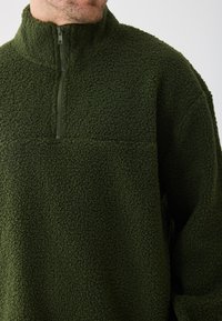 Grøn fleece pullover med høj krave, der har en halvdels lynlås. Struktureret, blødt materiale med afslappet pasform og uden synlige logoer.