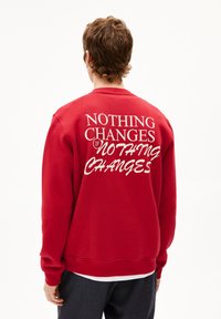 Rode sweatshirt van fleece, met op de achterkant witte tekst: "NIETS VERANDERT ALS NIETS VERANDERT." Geribbelde manchetten en zoom.