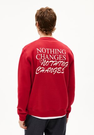 Rode sweatshirt van fleece, met op de achterkant witte tekst: "NIETS VERANDERT ALS NIETS VERANDERT." Geribbelde manchetten en zoom.