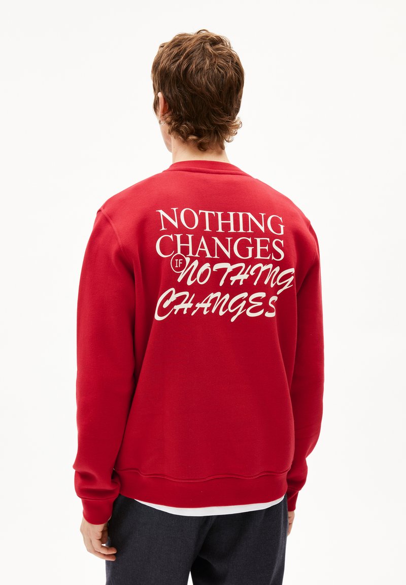Rode sweatshirt van fleece, met op de achterkant witte tekst: "NIETS VERANDERT ALS NIETS VERANDERT." Geribbelde manchetten en zoom.