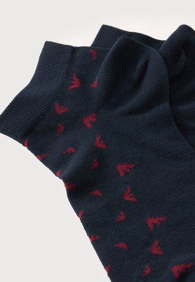 Paire de chaussettes bleu foncé avec petits motifs abstraits rouges en forme d’oiseaux et bordures côtelées, posées à plat sur fond blanc.