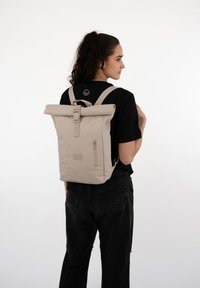 Beige roll-top backpack with adjustable straps, zip pocket, and logo detail. Made from durable fabric, features a clean, minimalist design.