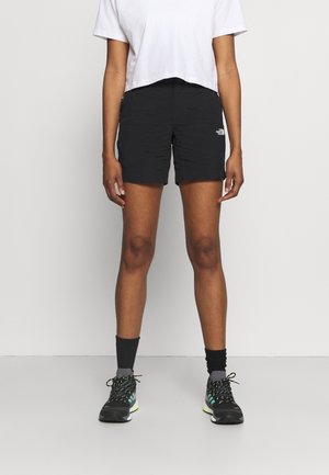 Personne portant un short noir North Face, un t-shirt court blanc, des chaussettes noires et des chaussures de sport noires, debout devant un fond uni.