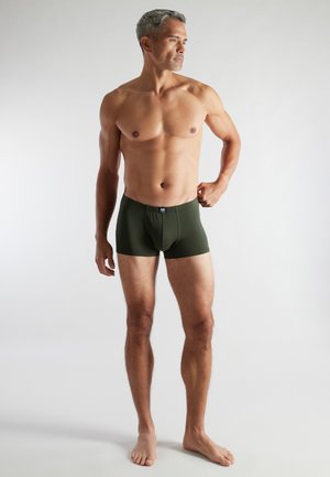 Ein grauhaariger Mann ohne Oberbekleidung steht barfuß in dunkelgrünen Boxershorts und blickt seitlich vor einem schlichten, hellen Hintergrund.