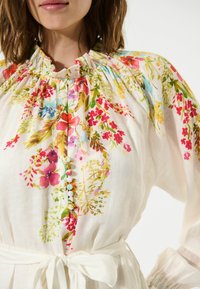 Blumenbluse aus leichtem, luftigem Stoff mit lebhaften Mustern in Pink, Blau und Grün. Elastischer Ausschnitt und Knopfdetails auf der Vorderseite.