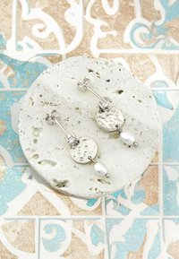 Söljearmband med en hamrad cirkulär design och en vit pärlaccent. De hänger från en enkel studpost på en texturerad bakgrund.