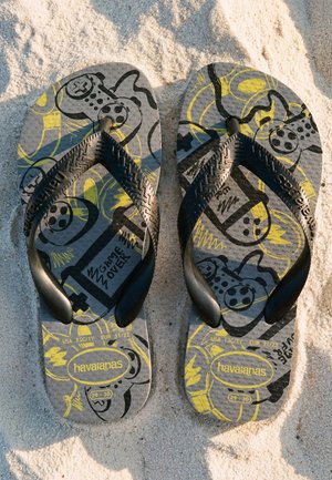 Paar schwarze und graue Havaianas Flip-Flops mit gelben und schwarzen Videospiel-Controller-Designs, die auf sandigem Boden liegen.