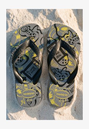 Paire de tongs Havaianas noires et grises avec des motifs de manette de jeu vidéo jaunes et noirs reposant sur un sol sablonneux.
