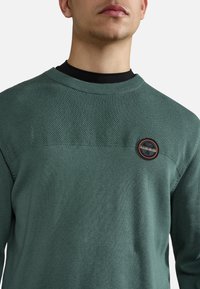 Groene gebreide trui met een textuur bovenpaneel, geribbelde halslijn en een ronde logo-patch op de borst. Glad stof voor het lichaam met een ontspannen pasvorm.