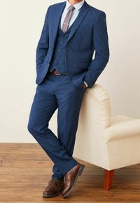 Marineblauwe driedelige maatpak met een notch lapel, een gestreepte das en bruine leren schoenen. Gladde stof en op maat gemaakt fit. Staande naast een stoel.