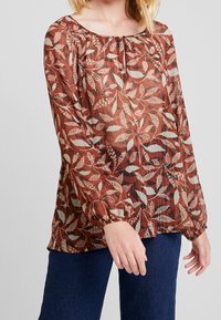 Blouse marron à motif de feuilles avec un col froncé et des manches longues légèrement bouffantes ; fabriquée en tissu léger et texturé.