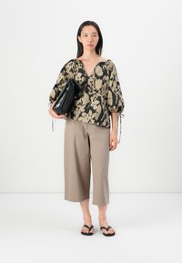 Blouse à motifs floraux, noir et beige avec des manches bouffantes, associée à un pantalon large beige et des tongs noires. Portant un sac noir.