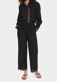 Svart prickig tvådelad outfit med en knapp-up blus och byxor med elastisk midja. Har scalloped kanter och vit dekorativ sömnad.