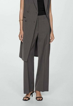 Femme portant un pantalon gris à jambes larges, un blazer sans manches assorti, un haut noir, un bracelet en or et des talons ouverts noirs, debout sur un fond uni.