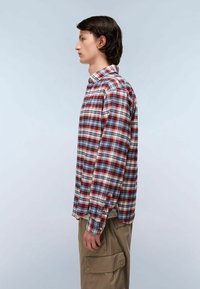 Giovane uomo in camicia a quadri rossa, bianca e blu e pantaloni cargo beige che sta in profilo contro uno sfondo chiaro e semplice.