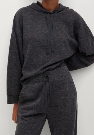 Personne portant un sweat à capuche en maille gris foncé avec des cordons de serrage et un pantalon assorti à cordons de serrage, debout avec une main sur la hanche.