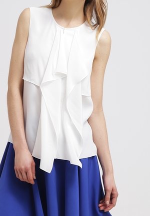 Blouse blanche sans manches avec détail à volants superposés, portée avec une jupe bleu roi mi-longue sur un fond neutre.