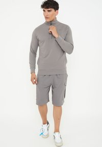 Grå atletisk zip-up topp med långa ärmar, kombinerad med matchande shorts med cargofickor, bärs med vita och grå sneakers.