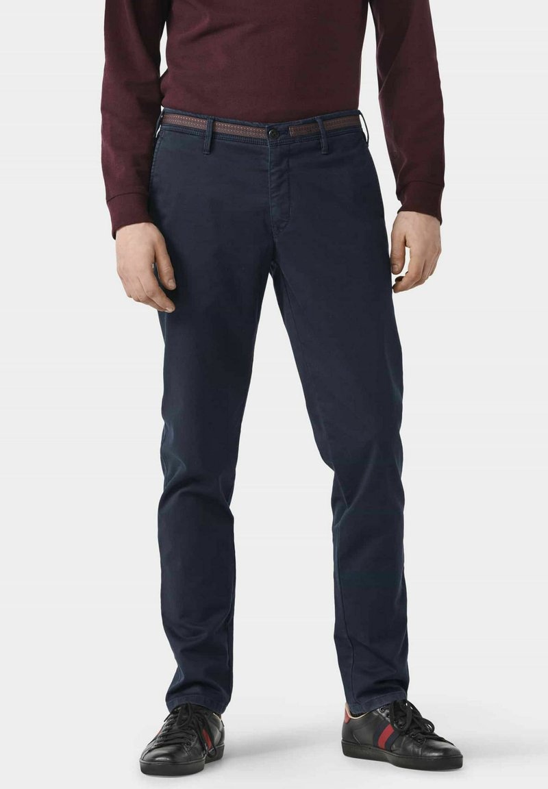 Marineblaue Baumwoll-Chinos mit leicht schmalerem Schnitt, kombiniert mit schwarzen Sneakers mit roten und grünen Akzenten sowie einem kastanienbraunen Langarmoberteil.