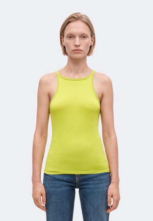 Une femme présente un haut sans manches jaune-vert vif et un jean bleu sur un fond clair uni.