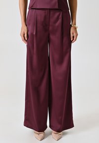 Pantaloni a gamba larga bordeaux in tessuto satin, con pieghe frontali e una texture liscia; abbinati a décolleté beige a punta.