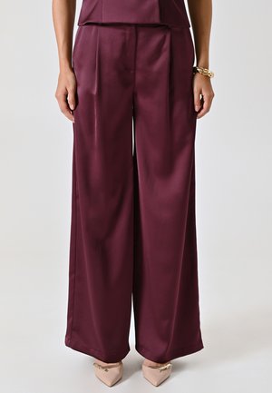 Pantaloni a gamba larga bordeaux in tessuto satin, con pieghe frontali e una texture liscia; abbinati a décolleté beige a punta.
