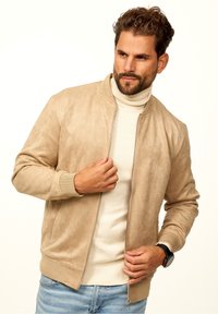 Beige suede bomber jacket with ribbed cuffs and waistband, zip closure, and side pockets, layered over a cream turtleneck sweater.