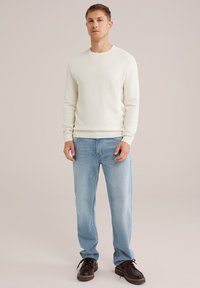 Cremfarbener Sweatshirt mit Textur, kombiniert mit hellblauen Jeans. Das Sweatshirt hat einen runden Halsausschnitt und gerippte Bündchen. Schwarze Schuhe runden das Outfit ab.