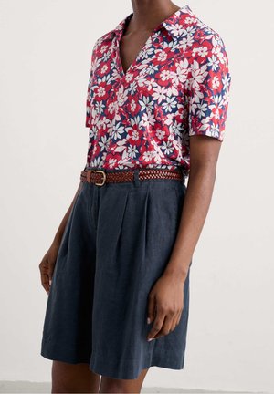 Personne portant une chemise à manches courtes rouge, blanche et bleue à motifs floraux, rentrée dans un short plissé bleu foncé taille haute avec une ceinture tressée marron.
