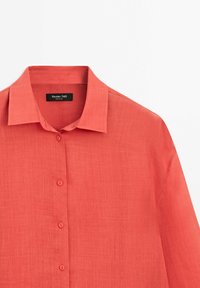 Chemise rouge corail à manches longues avec col classique et quatre boutons, tissu léger texturé.