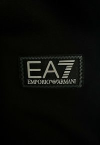Tessuto nero con un patch in gomma del logo EA7 Emporio Armani; presenta testo bianco e accenti leggermente in rilievo e texturizzati.