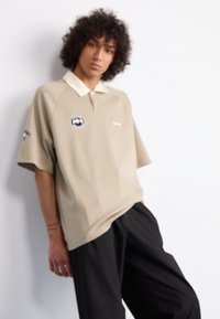 Beige polo shirt with a white collar, short sleeves, and a circular logo on the chest. Loose fit with soft texture; paired with black trousers.