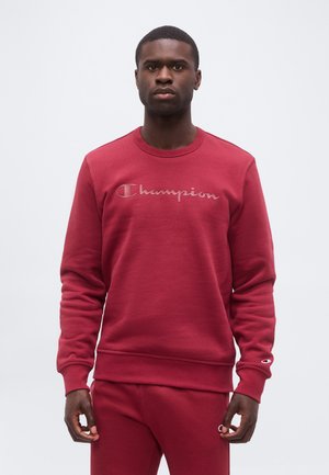 Homme portant un sweat-shirt et un pantalon rouge assortis de la marque Champion, debout devant un fond clair uni, face à l’avant avec une expression neutre.