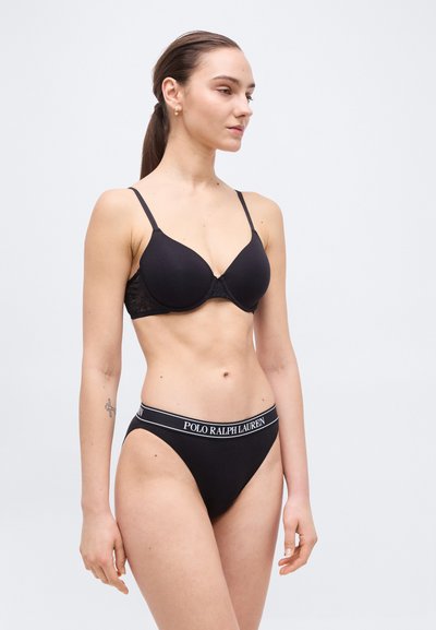 Γυναίκα σε μαύρο σουτιέν και μαύρα εσώρουχα Polo Ralph Lauren με λωρίδα μέσης με λογότυπο, κοιτάζοντας προς τα δεξιά σε απλό γκρι φόντο.
