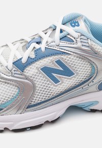 Primo piano di una sneaker New Balance bianca e azzurra chiara con tessuto a rete, sovrapposizioni sintetiche e lacci bianchi, che mette in evidenza il logo "N" sul lato.