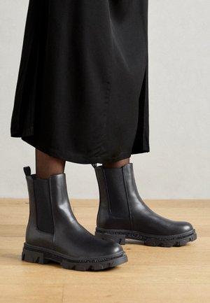 Czarne skórzane botki za kostkę z grubą, bieżnikowaną podeszwą, elastycznymi panelami po bokach i pętelkami do zakładania. Noszone z długim czarnym ubraniem, ukazując gładką fakturę.