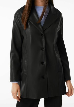 Kobieta ubrana w czarny, zapinany na guziki skórzany płaszcz na jasnoniebieskim swetrze, z rękami swobodnie opuszczonymi wzdłuż ciała na jednolitym białym tle.