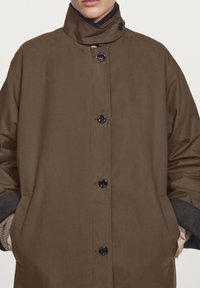 Brown outer coat featuring a high collar, button closure, and side pockets. The fabric appears smooth, with ribbed accents on the cuffs.