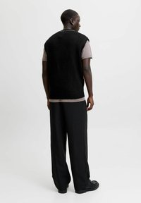 Man staat met zijn rug naar de camera, gekleed in een zwarte mouwloze trui over een beige T-shirt, zwarte gestreepte wijde broek en zwarte schoenen.