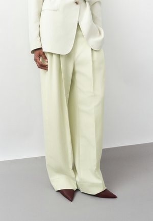 Osoba ubrana w kremowe spodnie o szerokich nogawkach i pasującą marynarkę, w połączeniu z ciemnobrązowymi, szpiczastymi butami, stojąca na tle jednolitego tła.