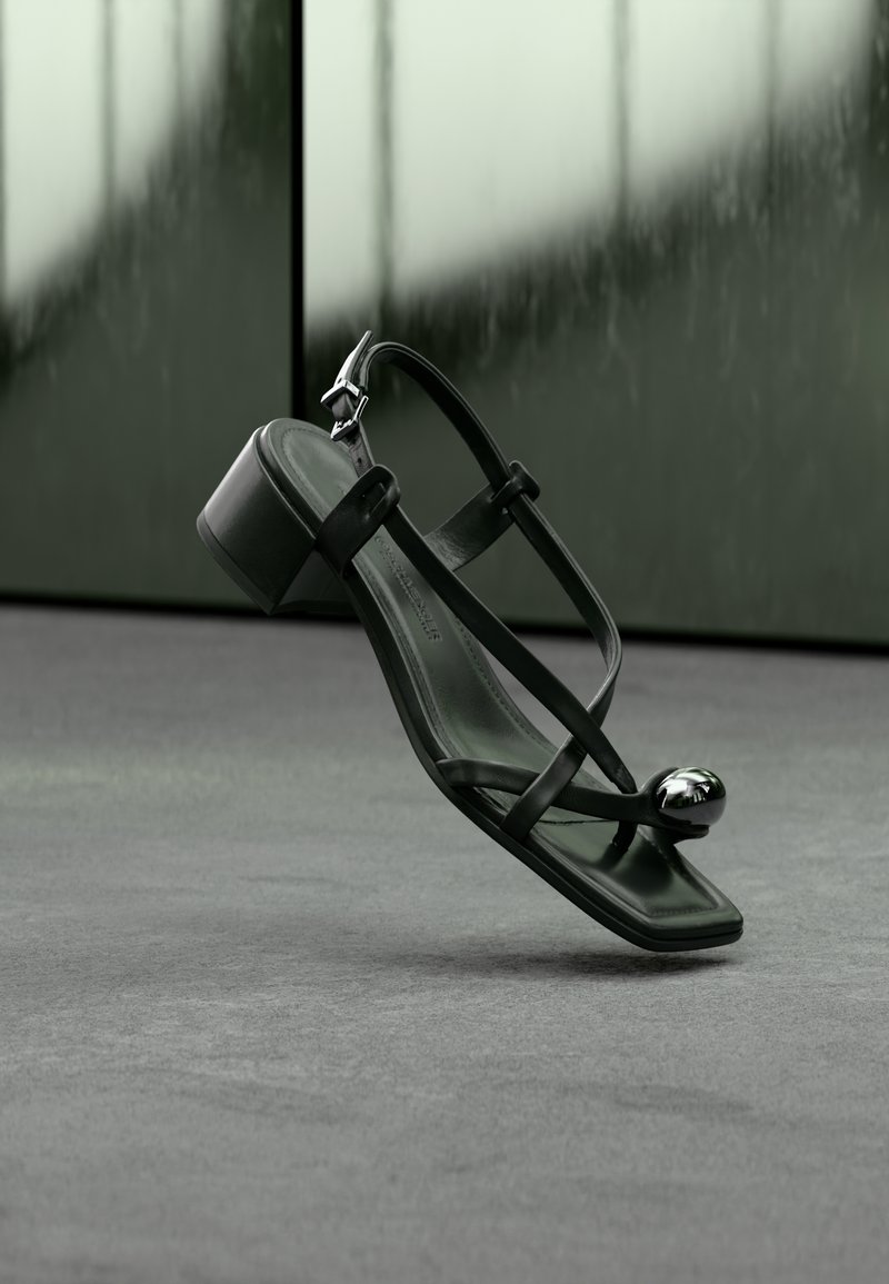 Sandalo nero con cinturini, tacco basso a blocco e perla decorativa argento sulla punta, mostrato sospeso sopra una superficie grigia con sfondo sfocato.