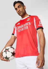 Rød fodboldtrøje med hvide detaljer, indeholdende "Emirates Fly Better" logo. Spilleren holder en bold med sort og hvidt mønster.